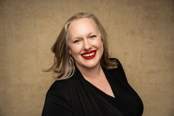 Elegant seated portrait of a woman with silver hair, wearing a sheer black dress adorned with intricate beading, posed confidently against a textured beige background.